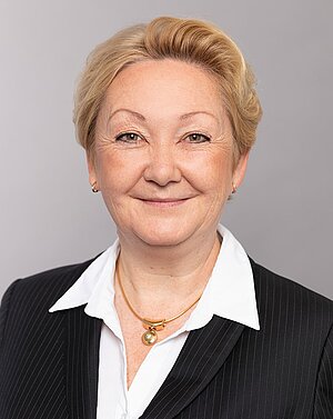 Porträt einer lächelnden Frau mit blonden Haaren, die einen schwarzen Anzug und eine weiße Bluse trägt. Sie hat eine goldene Halskette um den Hals. Der Hintergrund ist grau und neutral.