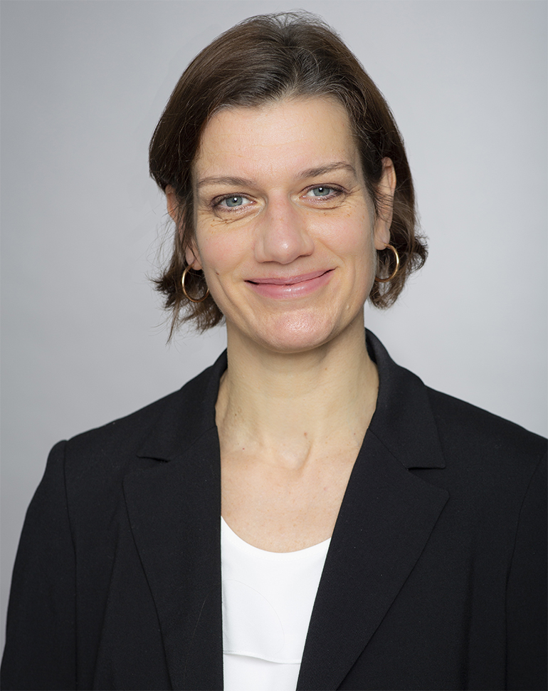 A woman with short brown hair and hoop earrings smiles warmly at the camera. She is wearing a black blazer over a white top, set against a neutral gray background. Her expression conveys confidence and approachability.