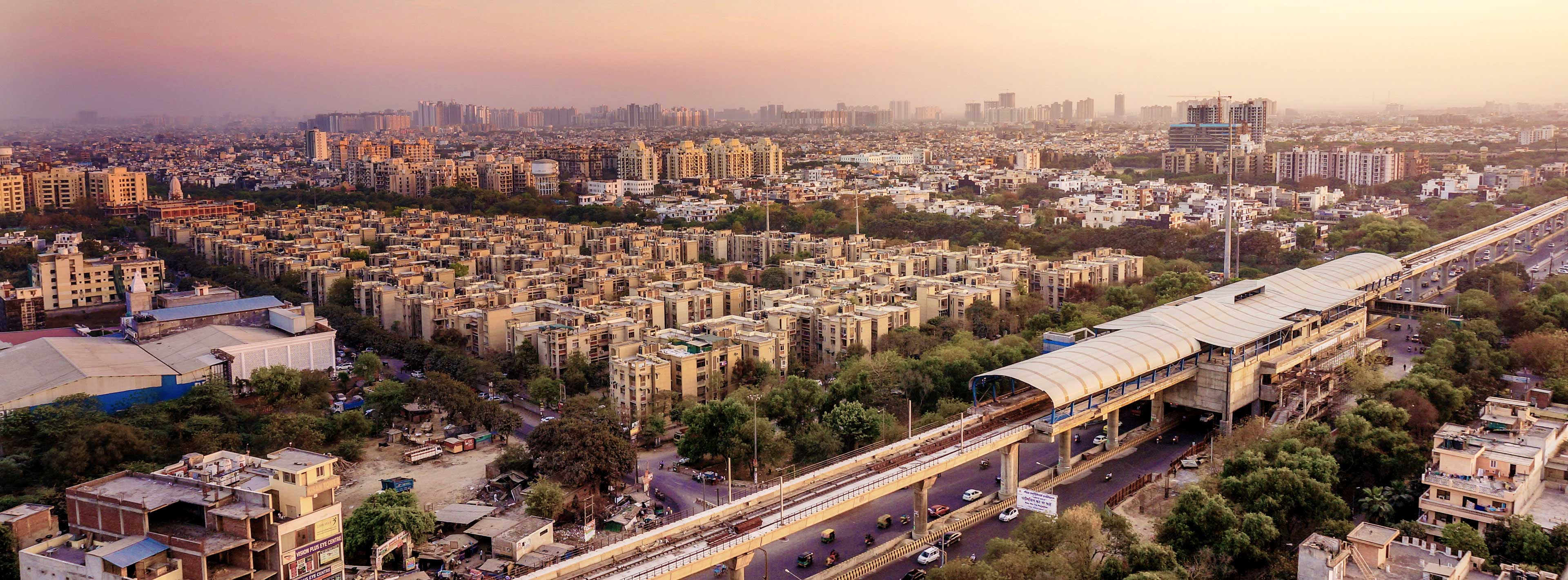 Aerial view of a densely populated urban area during sunset. Residential buildings with varying heights fill the landscape, interspersed with patches of greenery. A metro station and elevated train tracks are visible, indicating a developed transportation network in the city. The sky is softly lit with warm hues.