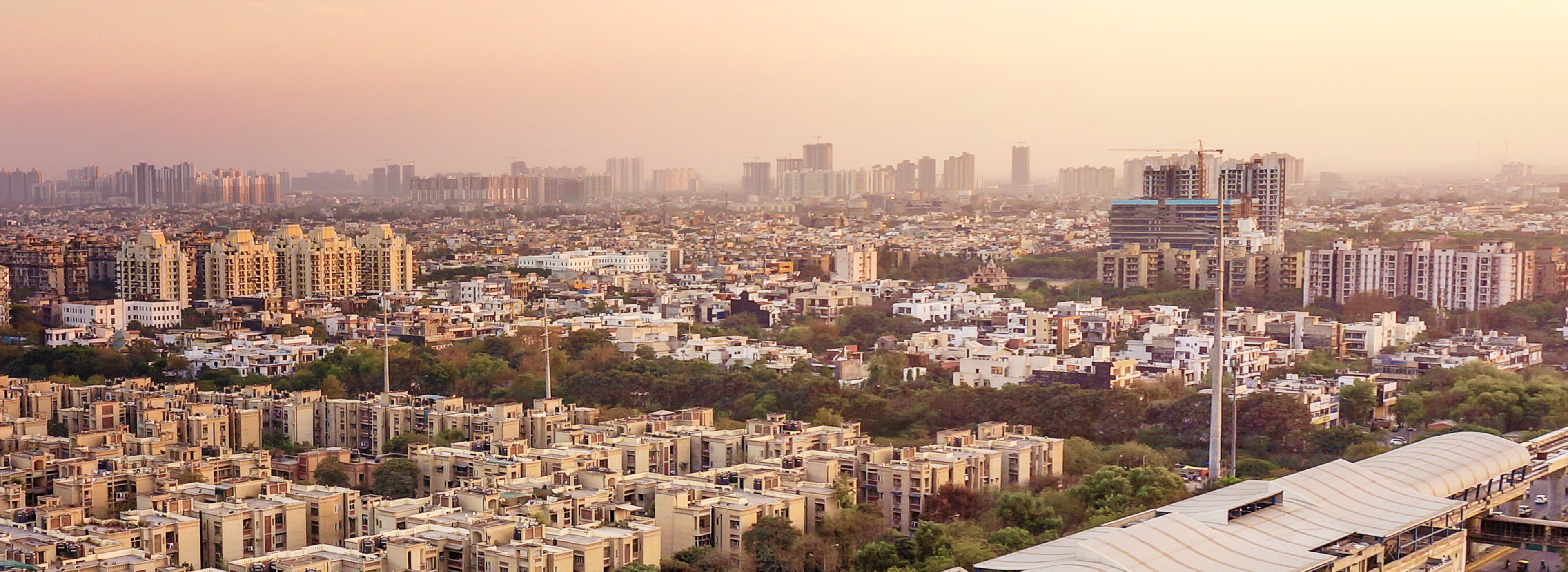 Luftaufnahme einer städtischen Landschaft mit zahlreichen Wohngebäuden und grünen Flächen. Im Hintergrund sind hohe Wolkenkratzer sichtbar, während das Licht eines Sonnenuntergangs den Himmel in sanften Farben erleuchtet. Die Szene vermittelt ein Gefühl von Urbanität und Natur im Einklang.