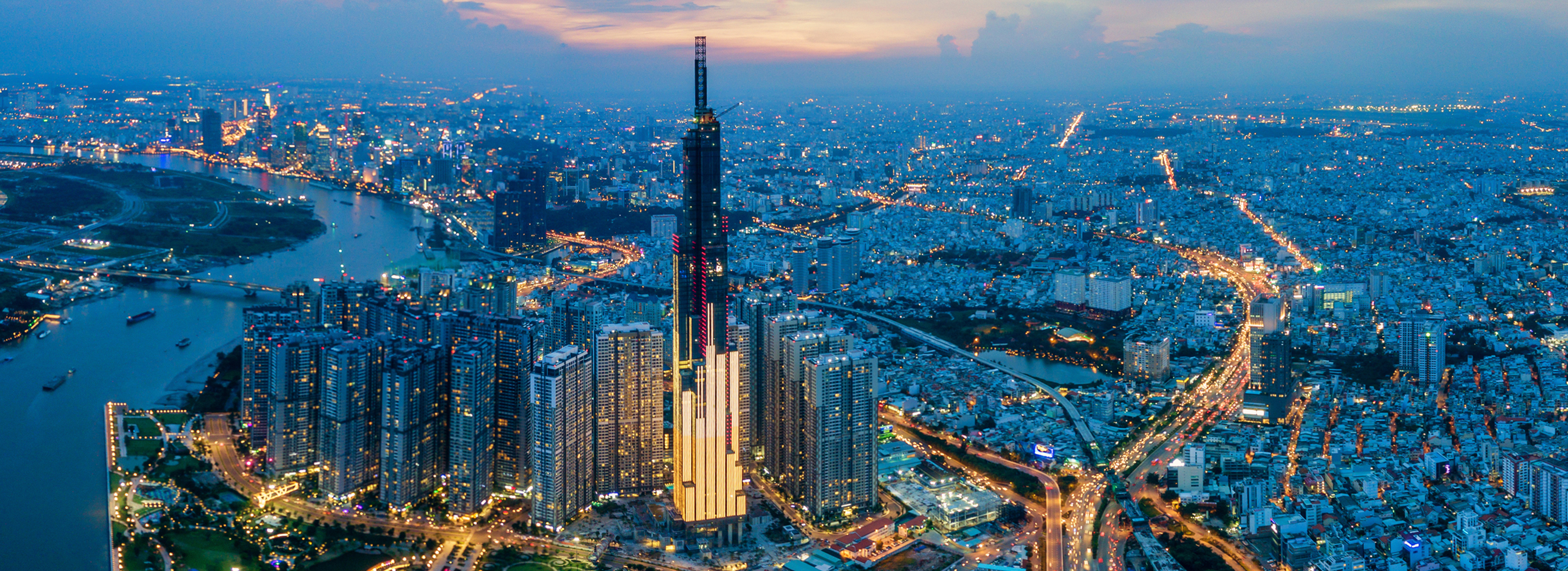Eine Luftaufnahme einer Stadt bei Dämmerung. Im Vordergrund steht ein hoher Wolkenkratzer, umgeben von weiteren Gebäuden. Lichter funkeln in den Straßen und am Wasser. Der Himmel ist leicht bewölkt und zeigt eine sanfte Farbpalette von Blau und Orange.