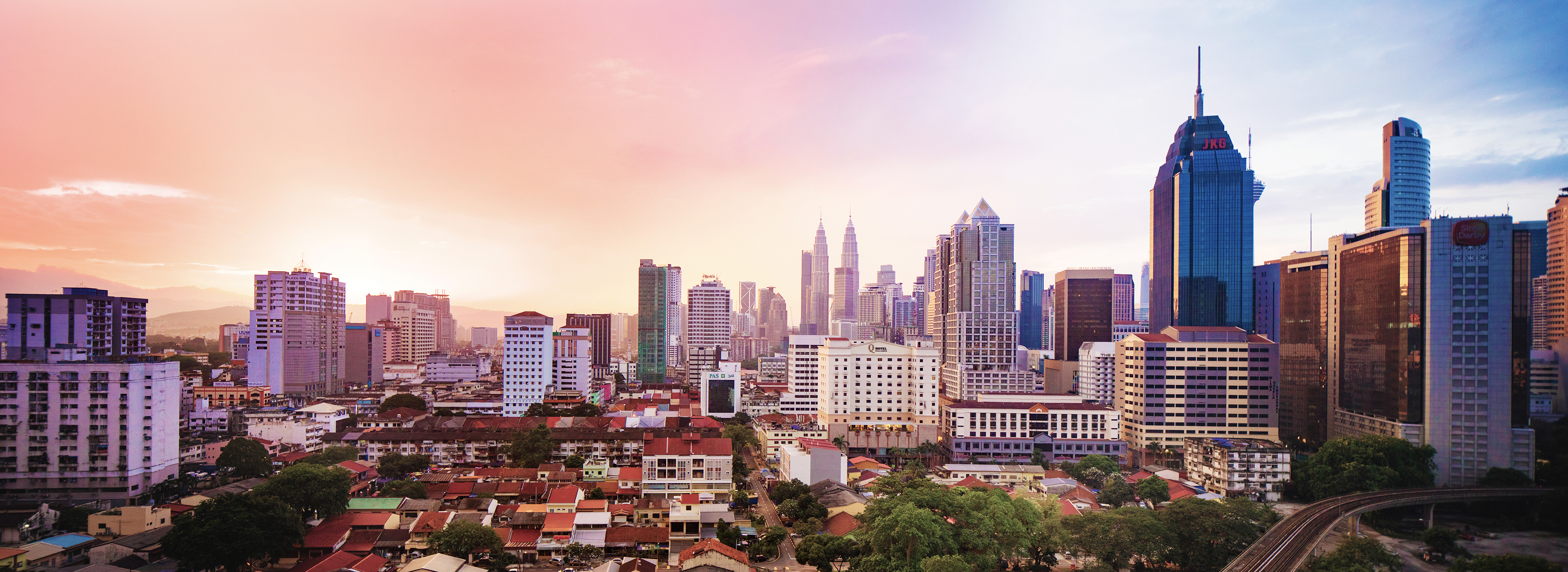 Die Skyline einer Stadt mit modernen Wolkenkratzern und einem sanften Sonnenuntergang im Hintergrund. Vorne sind niedrigere Gebäude und grüne Bäume zu sehen. Die Mischung aus Architektur und Natur schafft eine dynamische städtische Atmosphäre.