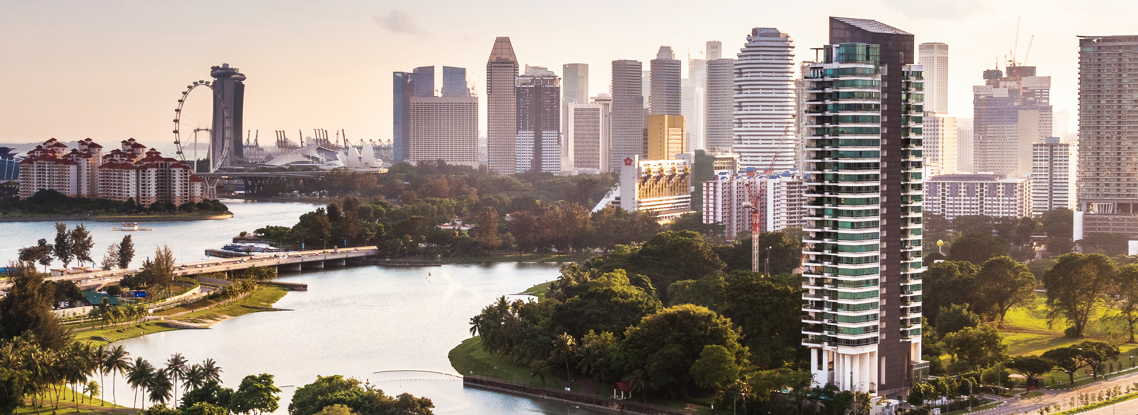 Blick auf eine moderne Stadtlandschaft mit hohen Gebäuden, umgeben von Grünflächen, einem Fluss und einer Promenade. Im Hintergrund ist ein Riesenrad und eine Uferpromenade zu sehen. Die Szene vermittelt eine Mischung aus Natur und urbanem Leben im frühen Morgenlicht.