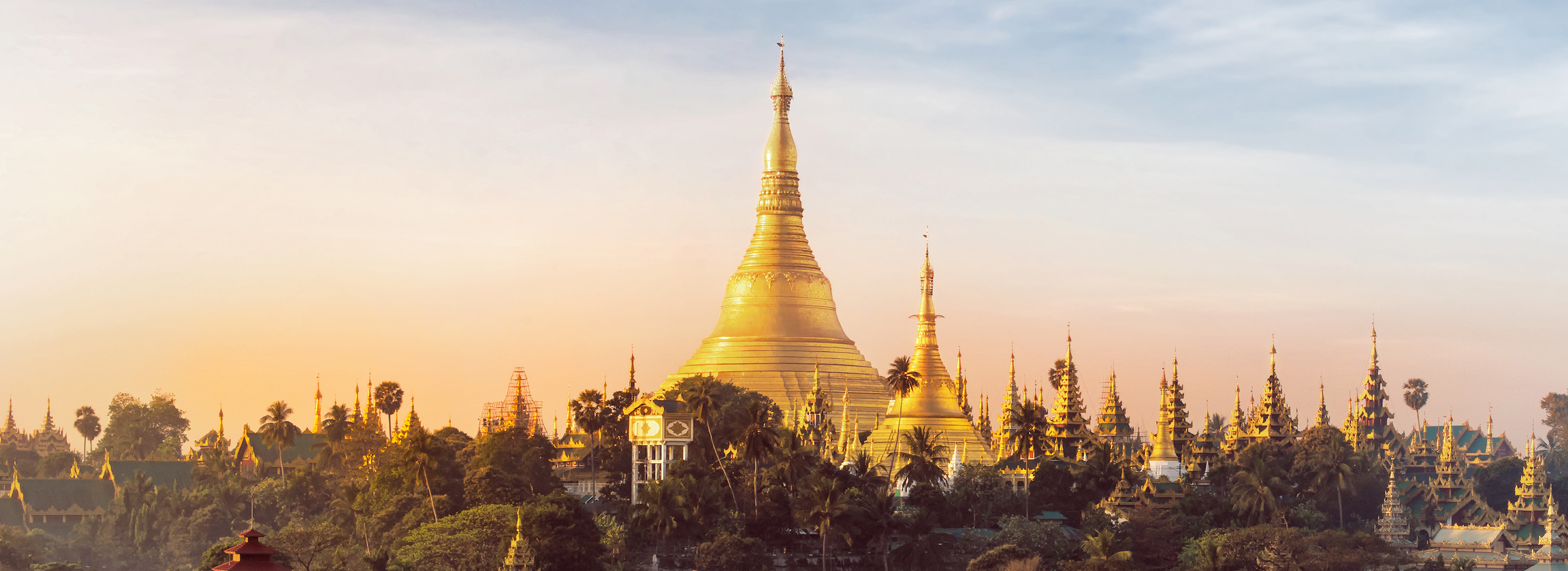 Eine goldene Stupa erhebt sich majestätisch vor einem sanften Sonnenuntergang. Umgeben von weiteren Pagoden und üppigem Grün, strahlt die Architektur in warmen Farben und vermittelt eine friedliche Atmosphäre. Die Szene symbolisiert die spirituelle und kulturelle Bedeutung des Ortes.