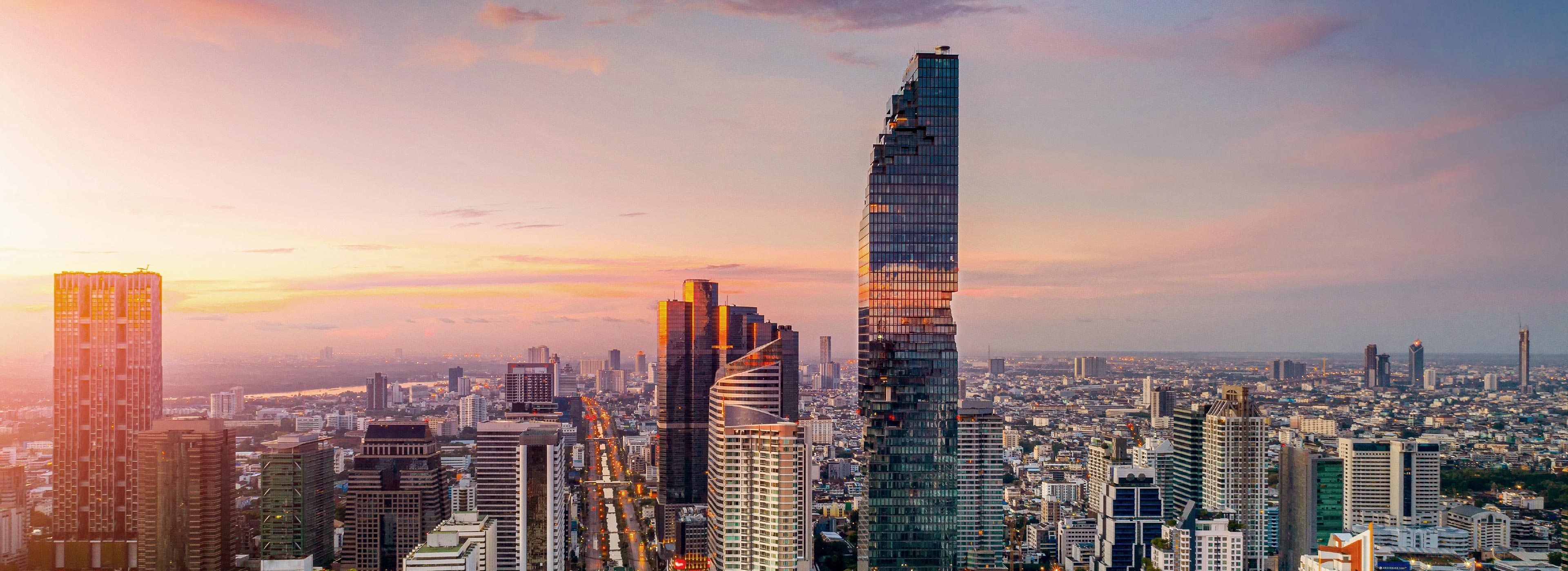 Eine atemberaubende Skyline einer Stadt bei Sonnenuntergang. Hohe, moderne Wolkenkratzer dominieren das Bild, während die sanften Farben des Himmels orange und lila Nuancen zeigen. Die Architektur ist vielfältig, mit reflektierenden Glasfassaden und einzigartigen Formen.