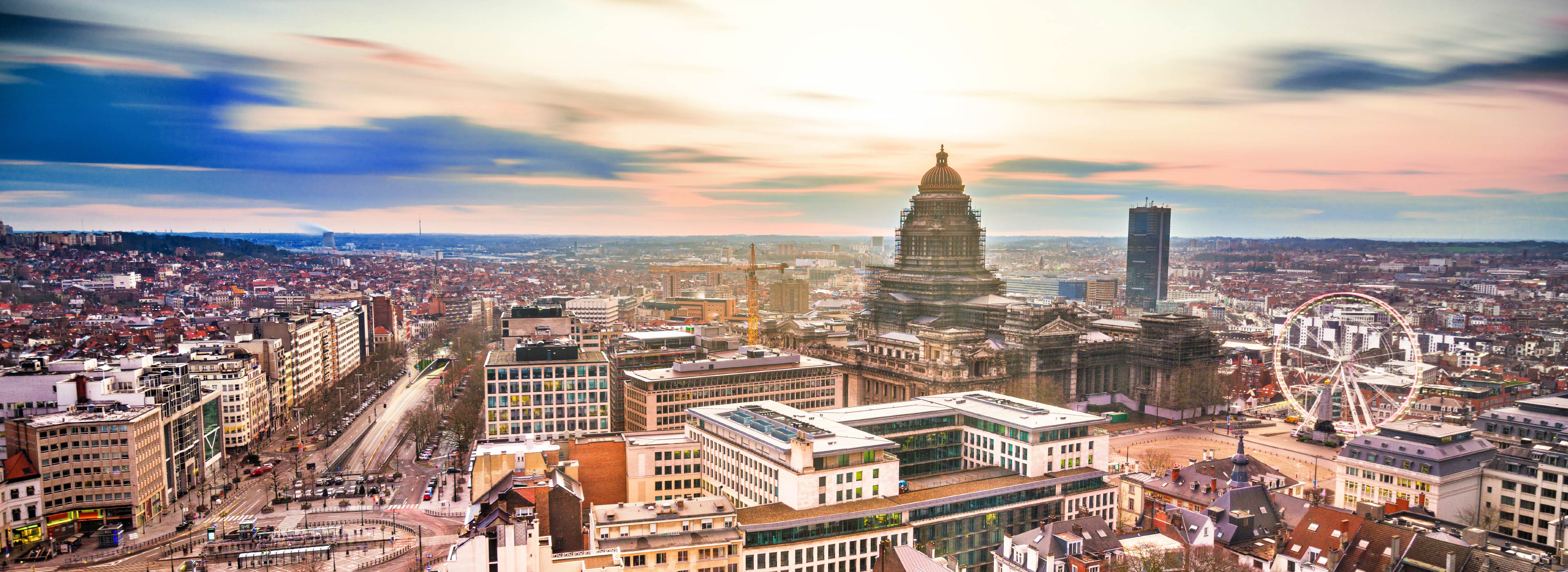 Eine weitläufige Stadtansicht zeigt moderne Gebäude und historische Architektur, darunter ein markantes, ornamentales Bauwerk im Vordergrund. Im Hintergrund ist ein Riesenrad zu sehen, umgeben von der urbanen Landschaft. Der Himmel ist farbenfroh bei Sonnenuntergang.