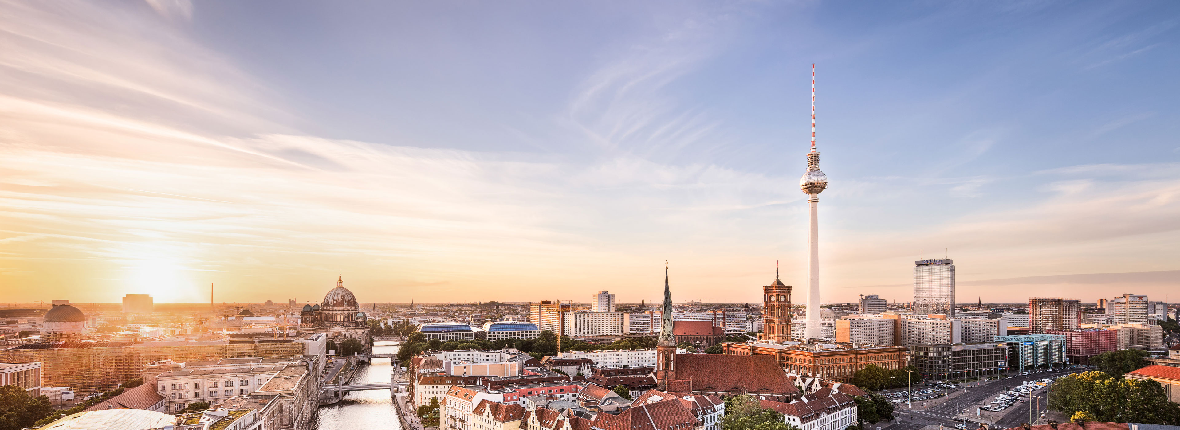 Panoramablick auf die Skyline von Berlin bei Sonnenuntergang. Im Vordergrund sind verschiedene Gebäude und die Spree zu sehen. Der Berliner Fernsehturm erhebt sich markant in die Höhe, während der Himmel in sanften Farben leuchtet.