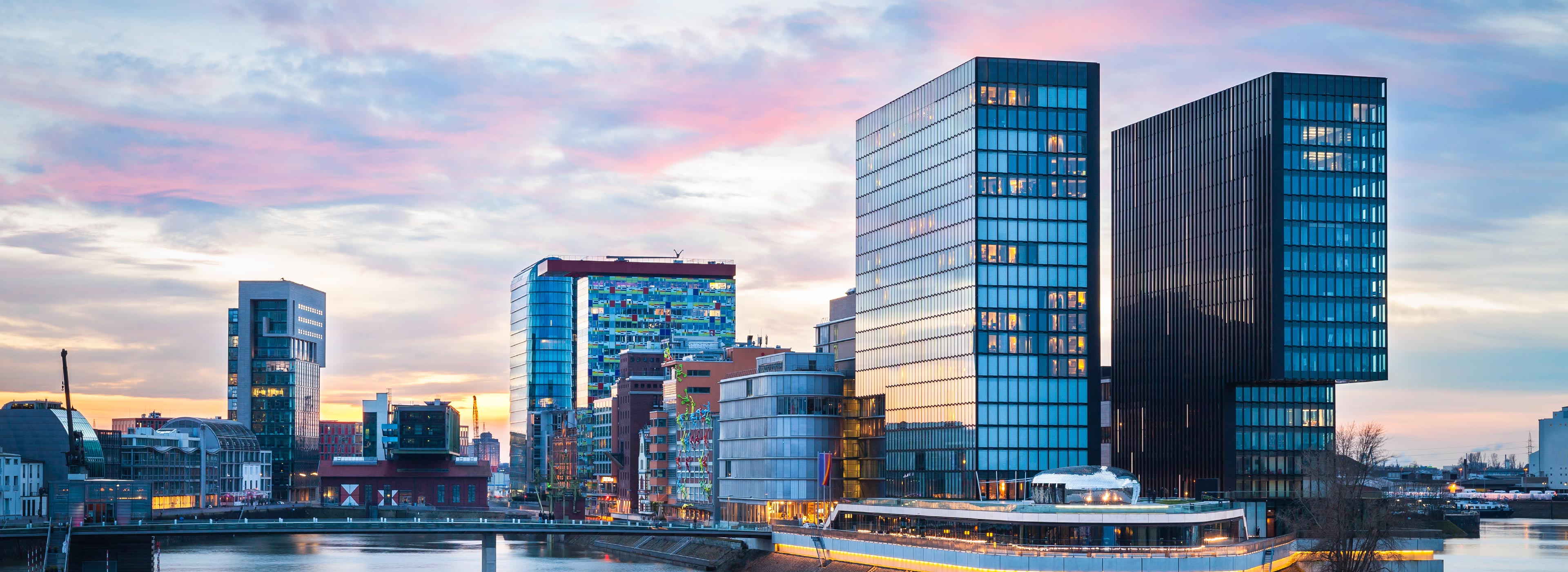 Städtische Skyline mit modernen Hochhäusern, deren Glasfassaden das Licht der Dämmerung reflektieren. Im Vordergrund verläuft ein Fluss, an dessen Ufer sich Restaurants und Cafés befinden. Der Himmel ist in sanften Farben wie Rosa und Blau gefärbt.