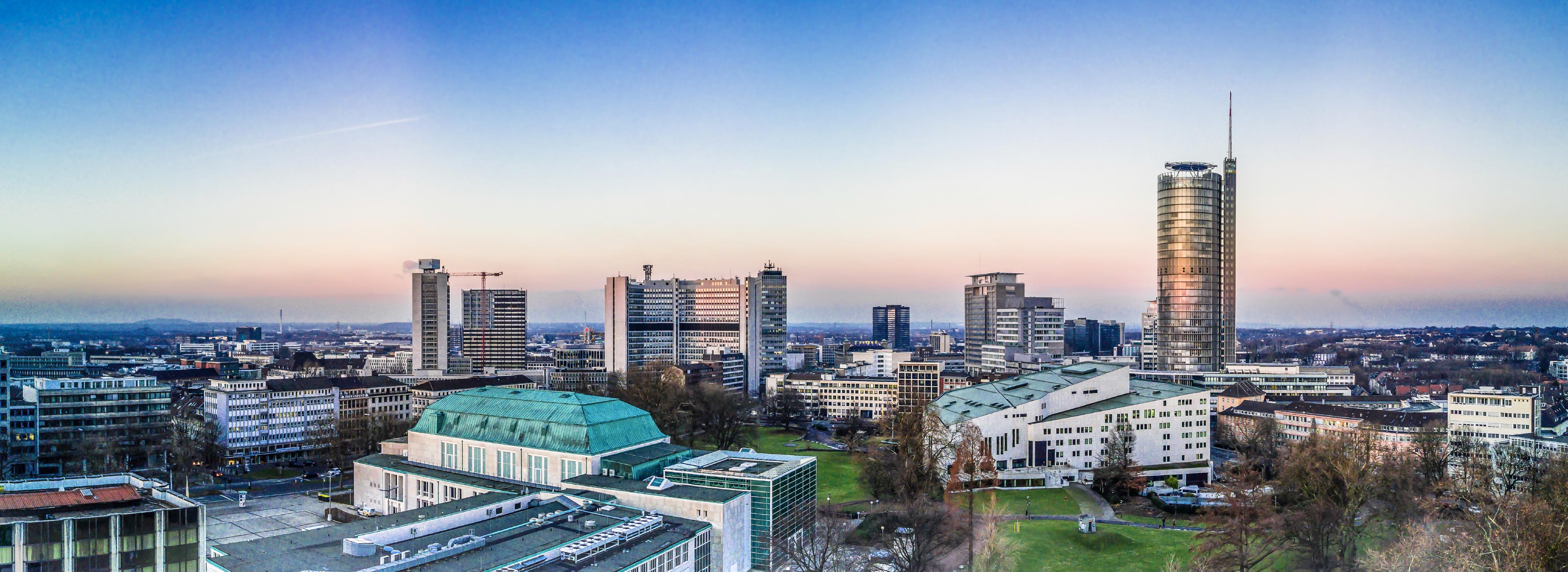Panoramablick auf eine moderne Stadtlandschaft mit hohen Gebäuden und einer grünen Parkfläche im Vordergrund. Der Himmel ist in sanften Farben der Dämmerung gefärbt, was eine ruhige Stimmung erzeugt.