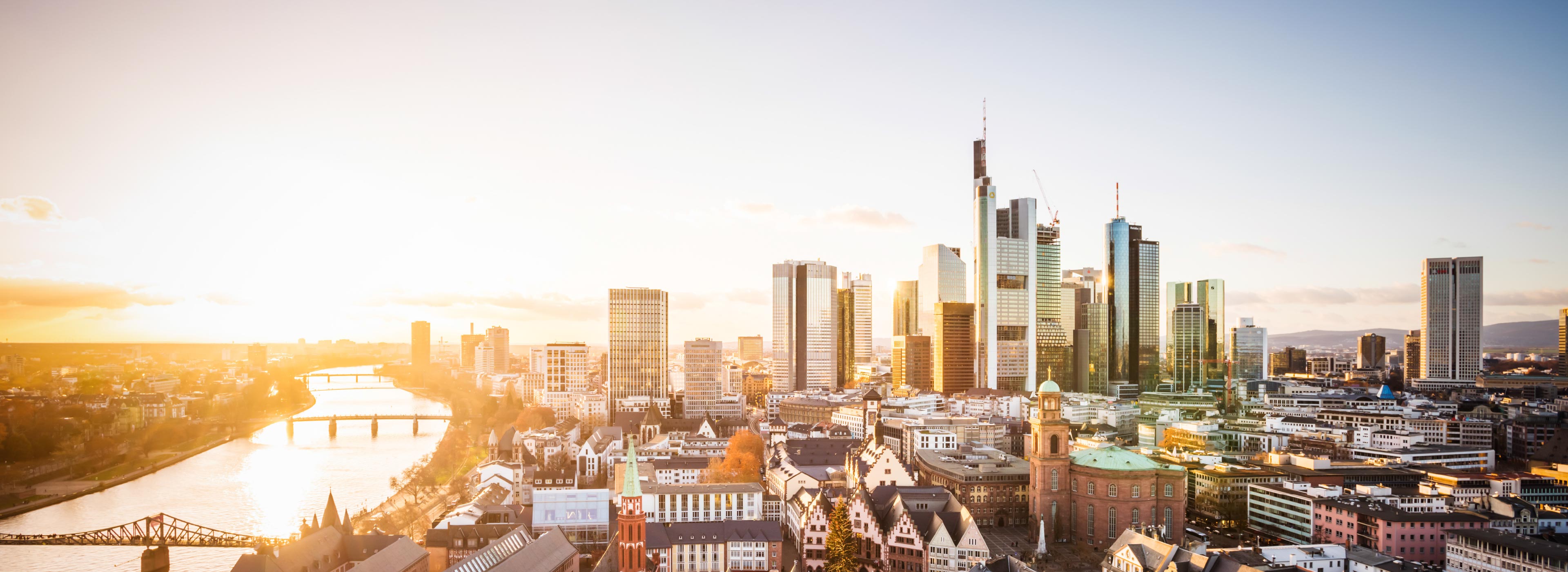 Eine Panoramaansicht der Skyline von Frankfurt am Main bei Sonnenuntergang. Hohe Wolkenkratzer und moderne Gebäude dominieren das Stadtbild, während die Sonne warmes Licht über die Stadt strahlt. Im Vordergrund sind einige historische Gebäude sowie der Fluss Main sichtbar.