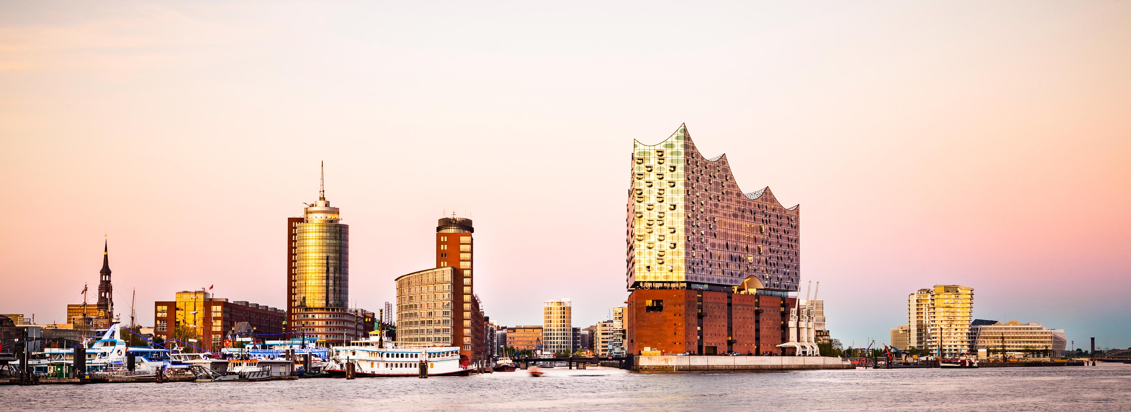 Eine Panoramaansicht des Hamburger Hafens bei Sonnenuntergang. Im Vordergrund sind mehrere Boote im Wasser zu sehen, während sich im Hintergrund moderne Gebäude, darunter die auffällige Elbphilharmonie, erheben. Der Himmel zeigt zarte Rosa- und Orangetöne.