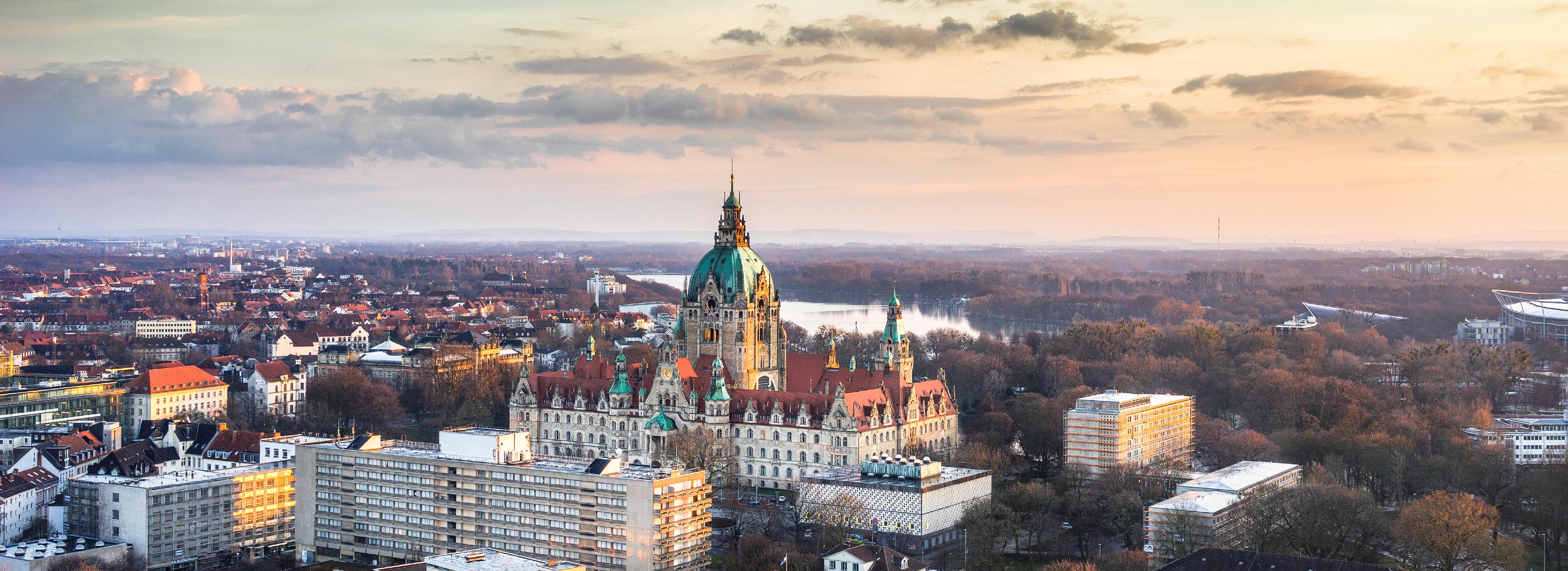 Blick auf die Stadt Hannover bei Sonnenuntergang. Im Vordergrund steht das historische Rathaus mit grünen Türmen und roten Dächern. Umgeben von einer Mischung aus modernen Gebäuden und Bäumen erstreckt sich die Stadtlandschaft bis zum Horizont.