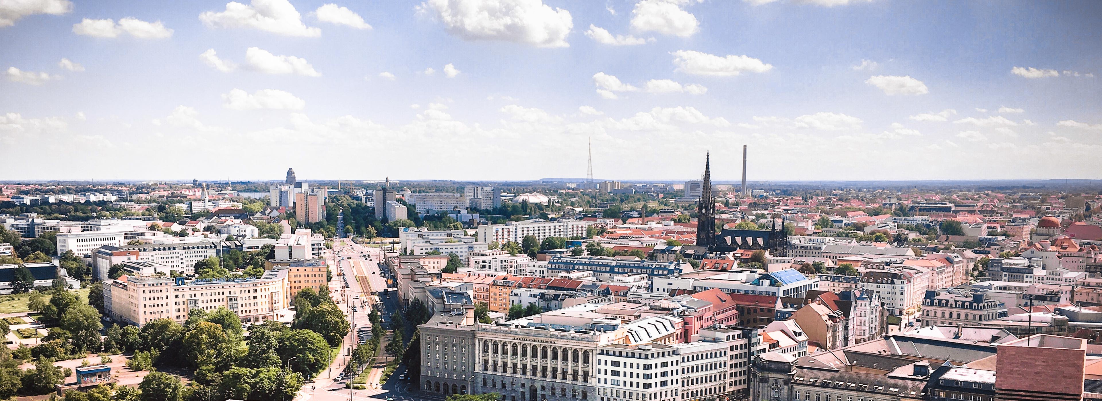 Eine weitläufige Stadtansicht zeigt eine Vielzahl von Gebäuden und Straßen unter einem klaren Himmel mit einigen Wolken. Im Vordergrund sind moderne und historische Bauwerke zu sehen, während im Hintergrund hohe Türme und Grünflächen vorhanden sind, die zur urbanen Landschaft beitragen.