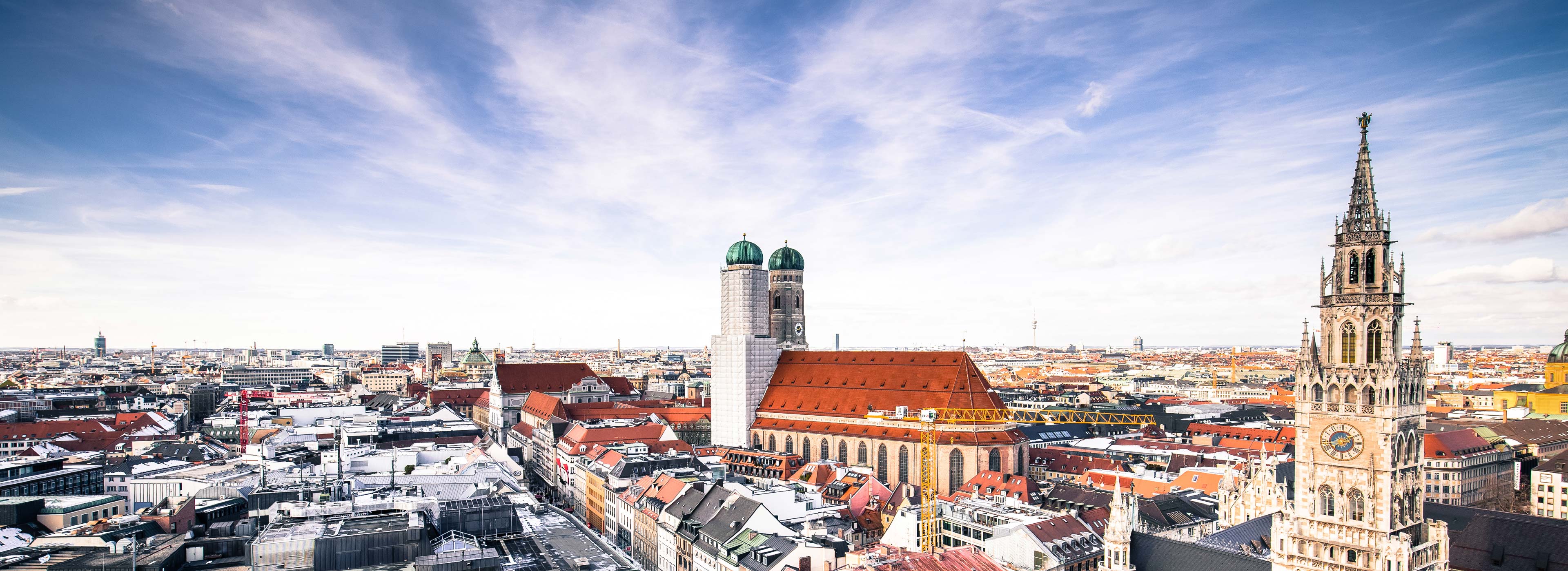 Eine Panoramaansicht von München zeigt die markanten Türme der Frauenkirche und das Neue Rathaus. Die Stadt ist von einer Vielzahl von Dächern umgeben, die im Sonnenlicht schimmern, und der Himmel ist hell und wolkenlos.