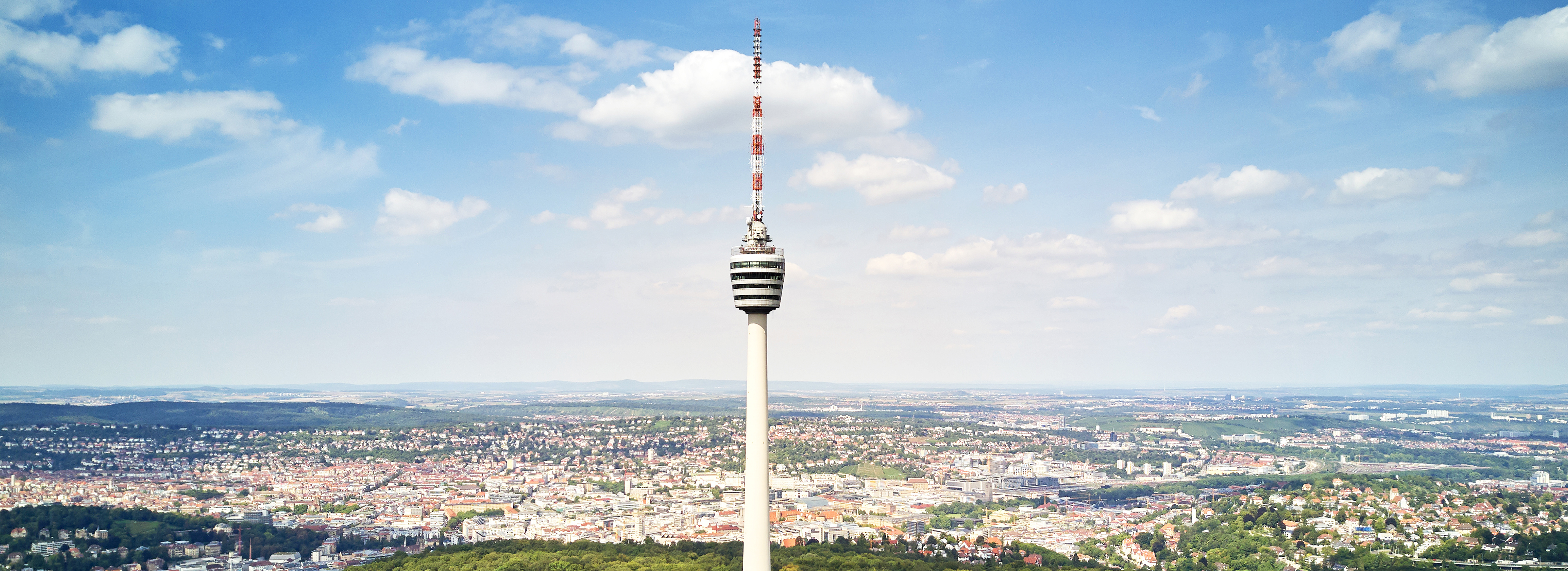 Der Fernsehturm erhebt sich hoch über einer Stadtlandschaft, umgeben von sanften Hügeln und unter einem weiten, blauen Himmel mit weißen Wolken. Er symbolisiert eine Aussicht auf die Umgebung und die städtische Architektur.