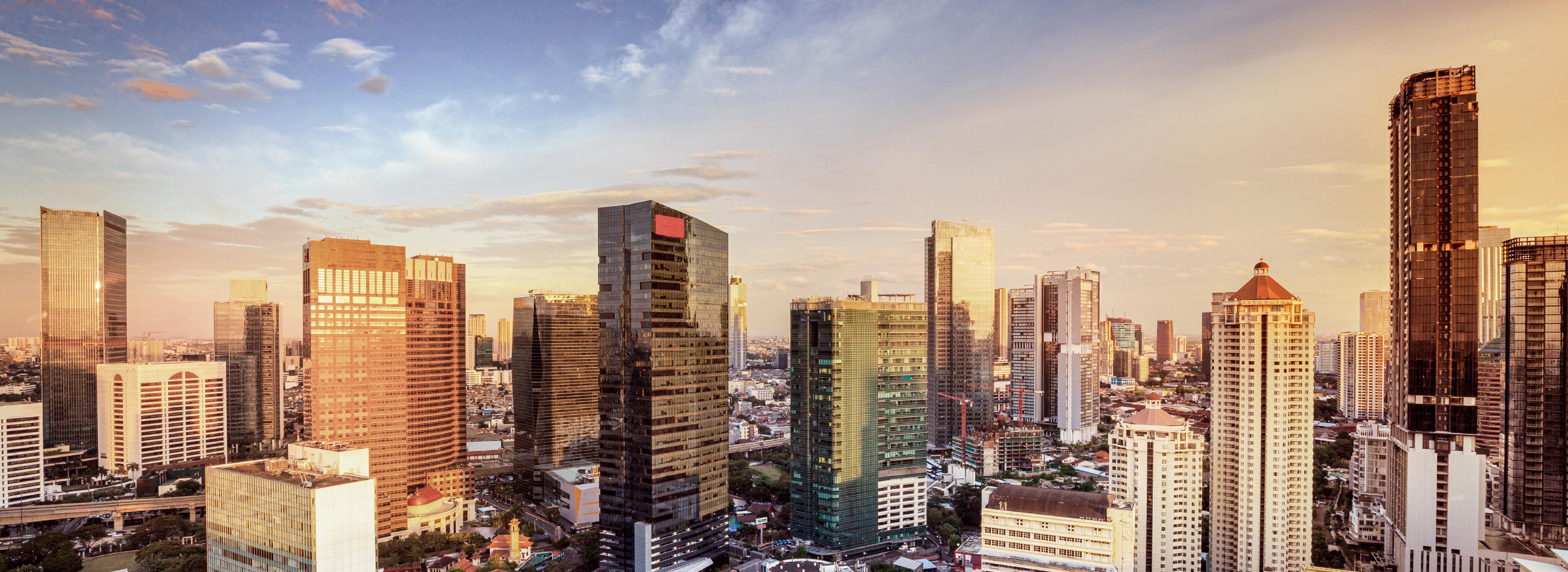 Eine Panoramaansicht einer modernen Stadt mit hohen Glas- und Stahlgebäuden, die im Licht der untergehenden Sonne funkeln. Der Himmel ist in sanften Farben gefärbt, während Straßen und Grünflächen sichtbar sind.