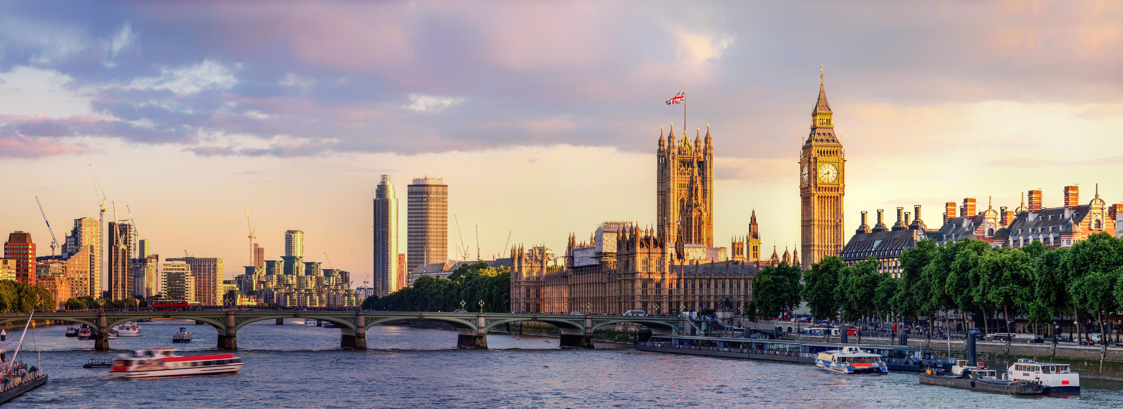 Panoramablick auf die Themse in London bei Sonnenuntergang. Im Vordergrund sind Boote auf dem Wasser zu sehen, während im Hintergrund das beeindruckende Parlamentsgebäude und der Big Ben zu erkennen sind. Die Szene vermittelt eine ruhige, stimmungsvolle Atmosphäre.