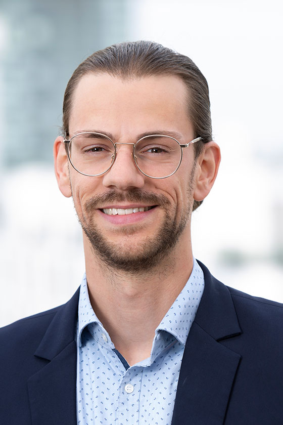 Porträt eines lächelnden Mannes mit Brille, der einen dunkelblauen Anzug und ein helles, gepunktetes Hemd trägt. Der Hintergrund ist unscharf, zeigt jedoch städtische Elemente. Der Mann wirkt freundlich und professionell.