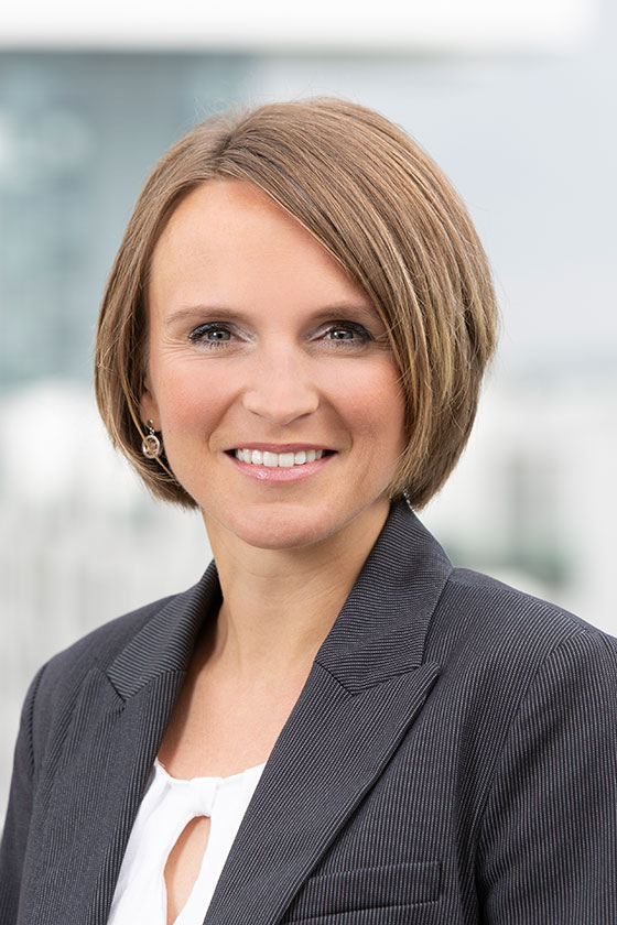 Porträt einer Frau mit schulterlangen, hellbraunen Haaren und einem freundlichen Lächeln. Sie trägt einen dunklen Blazer und eine helle Bluse. Der Hintergrund ist unscharf und zeigt eine moderne Büro-Umgebung.