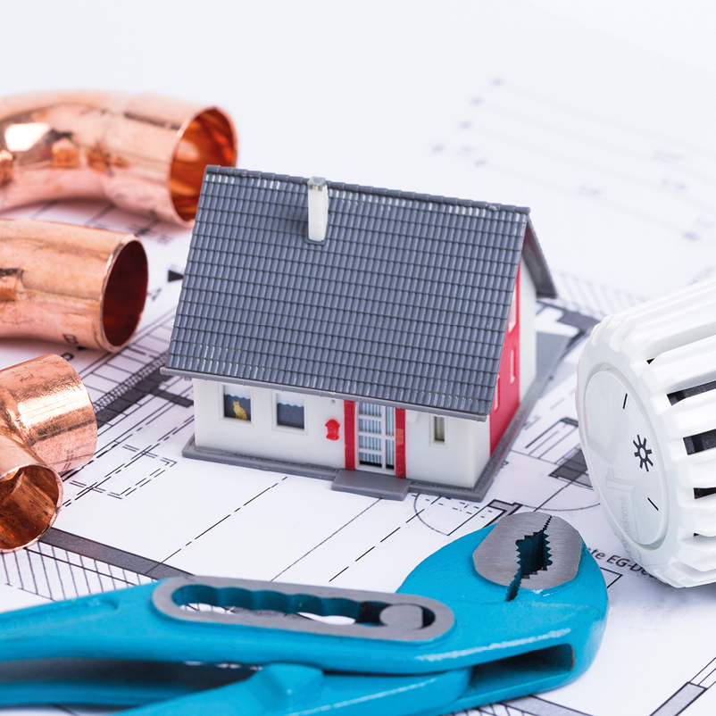 Ein Miniaturhaus steht auf Bauplänen, umgeben von Kupferrohren und einer Heizkörperthermostat. Dazu liegt eine Rohrzange. Die Anordnung deutet auf Installation oder Reparatur von Sanitär- und Heizungsanlagen hin.