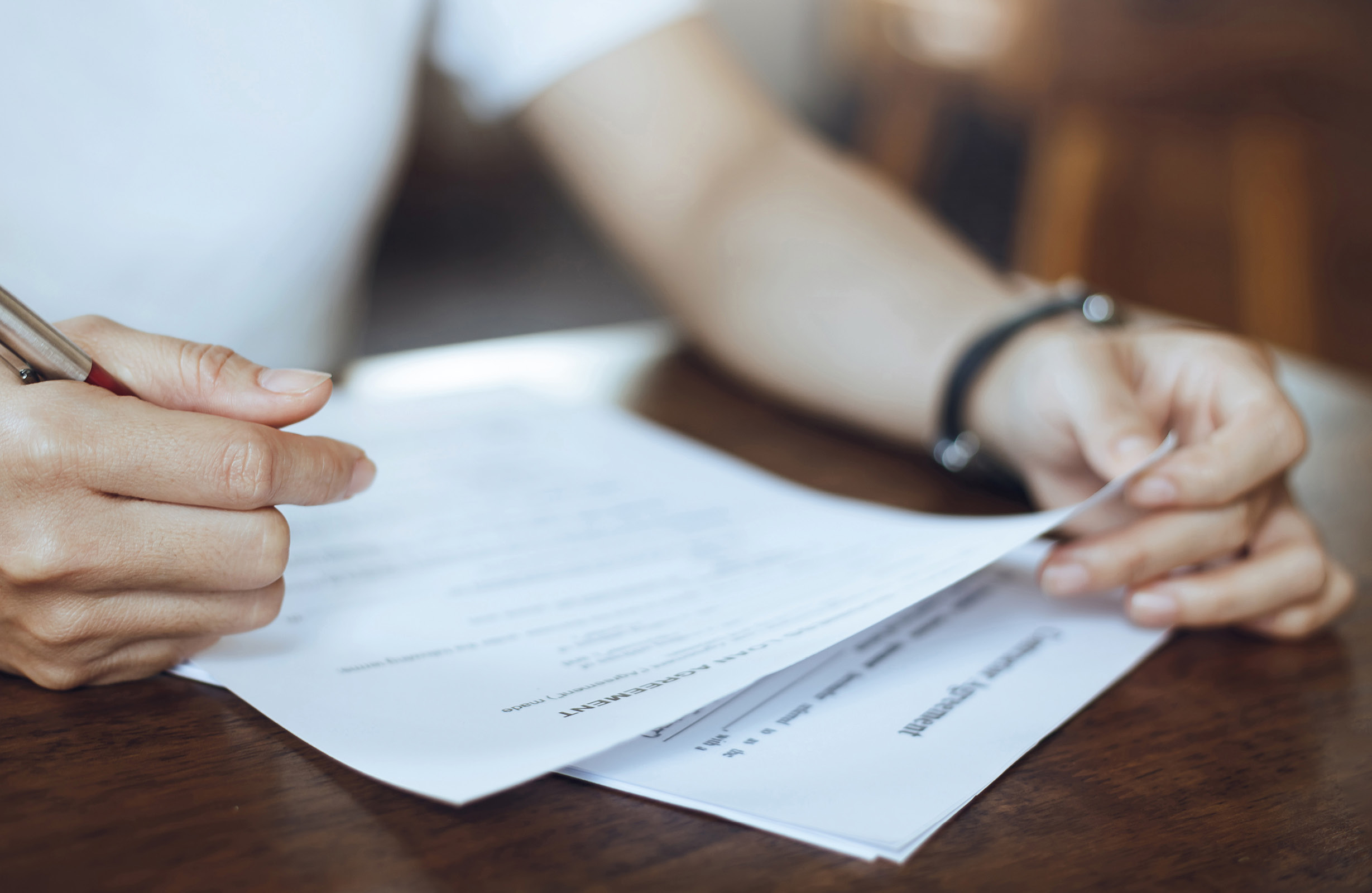 Eine Person hält einen Stift in der Hand und schaut auf mehrere Dokumente, die auf einem Tisch liegen. Die Hände sind im Fokus, während der Hintergrund unscharf ist.