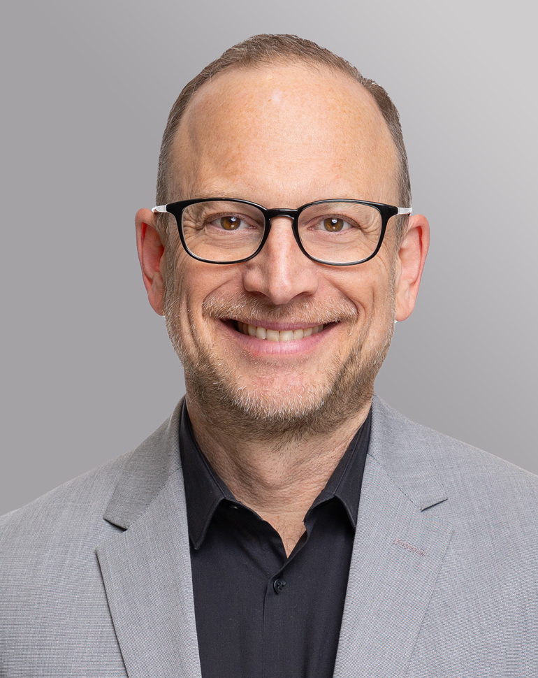 A smiling middle-aged man with short hair and glasses is wearing a gray blazer over a black shirt. The background is a soft gradient from light to dark gray.